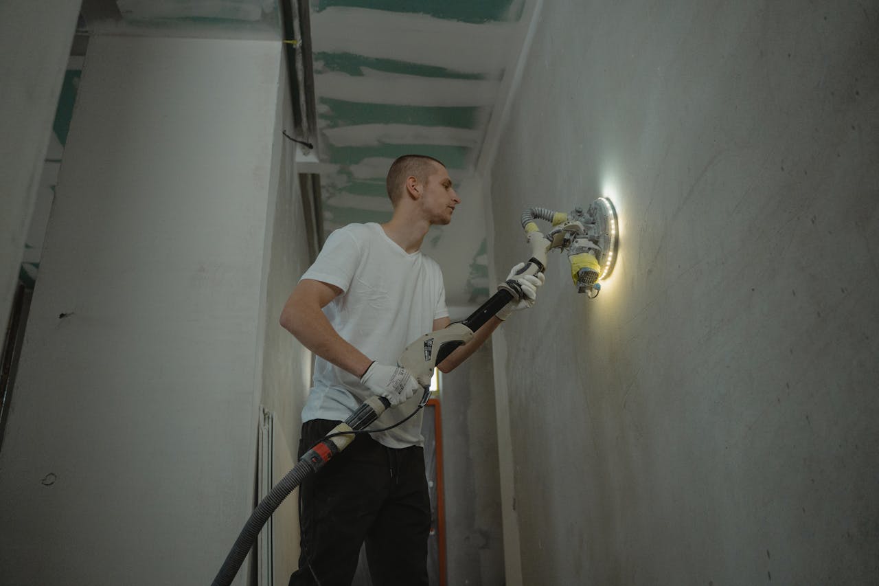 our-story A man sanding an interior wall with an automatic sanding tool during renovation work indoors.
